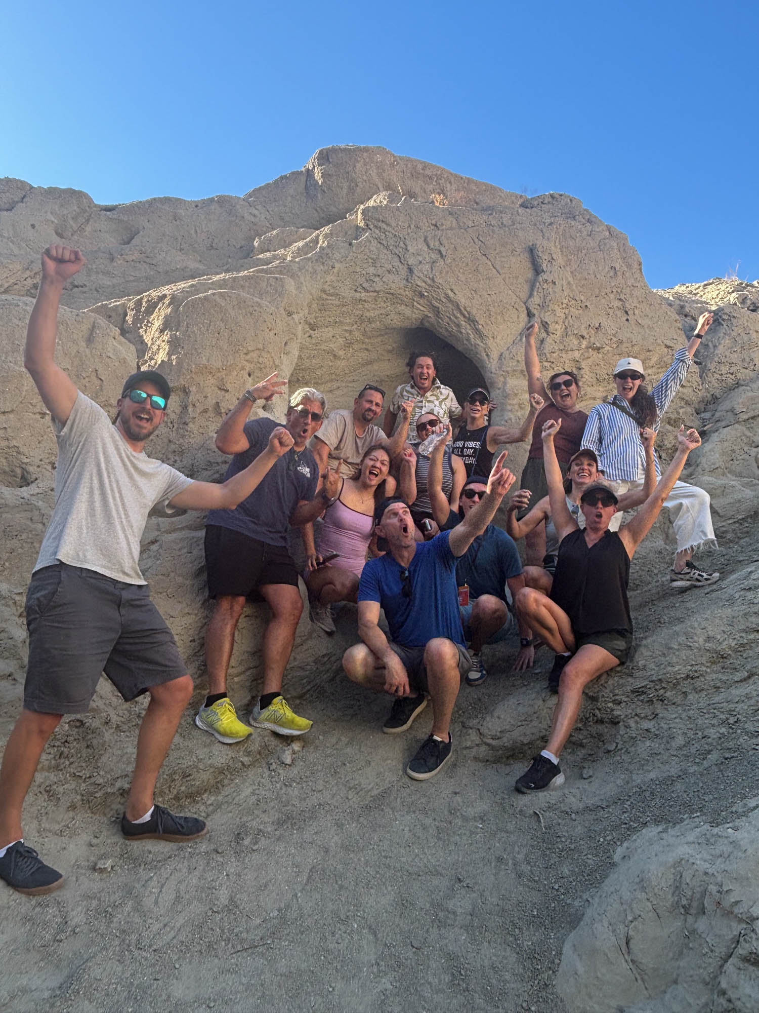 A group of people pose energetically on rocky terrain in front of a cave, with blue sky overhead. Some have arms raised and all appear to be enjoying the outdoor setting.