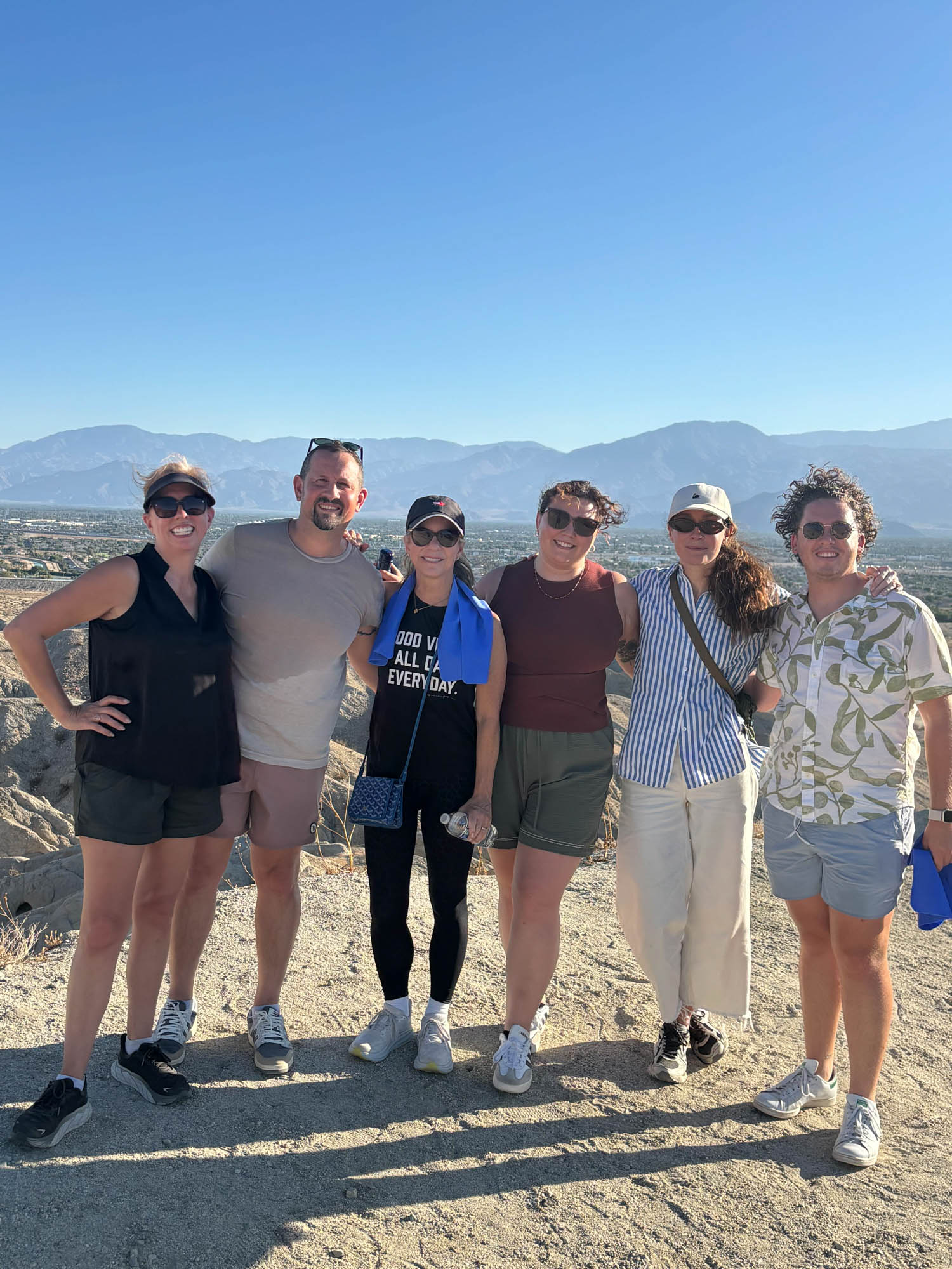 Six people stand together outdoors on a sunny day with mountains and blue sky in the background, dressed in casual summer clothing.