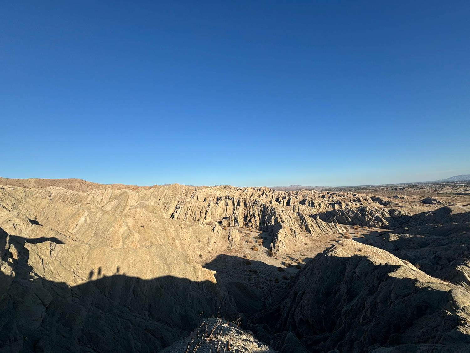 Desert badlands under a clear blue sky with rugged, eroded hills and deep shadows cast across the rocky landscape.
