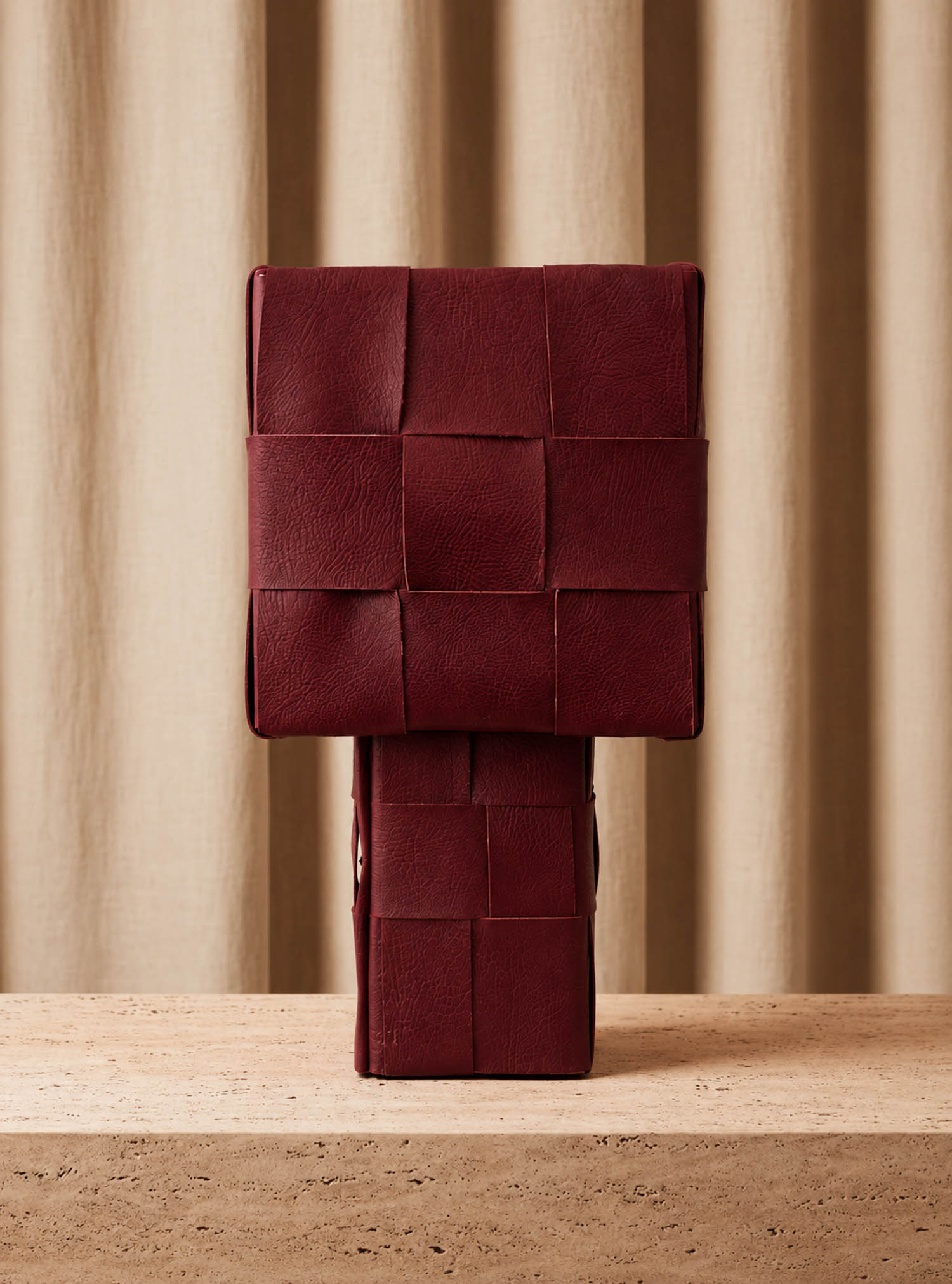 A rectangular, dark red leather handbag with a woven pattern displayed upright on a beige stone surface against a beige curtain background.
