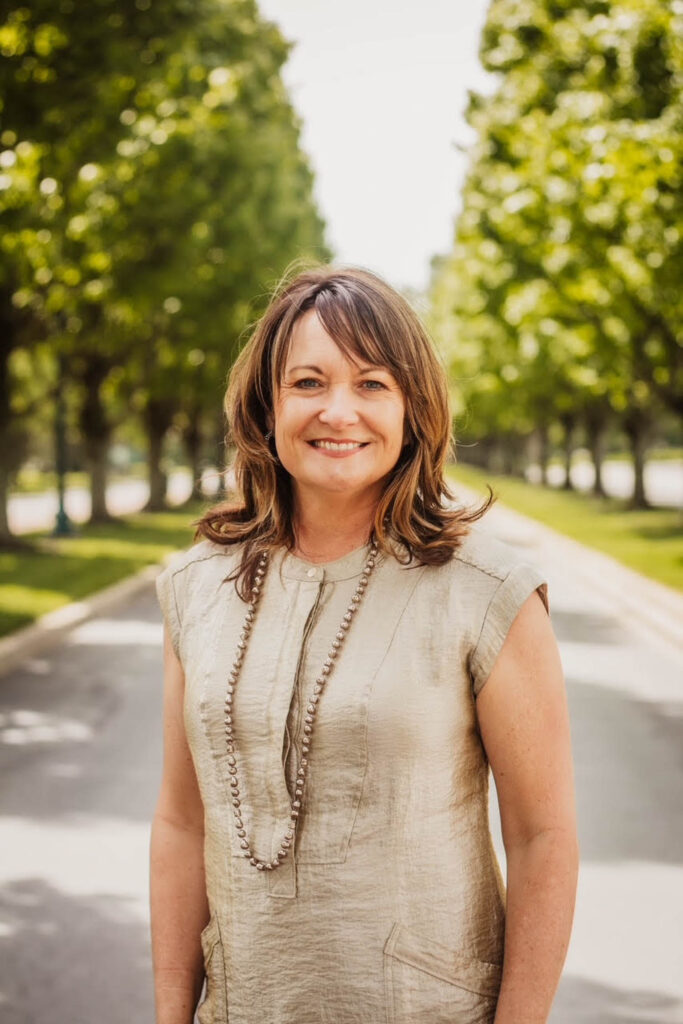 A woman in a beige dress stands smiling on a tree-lined pathway outdoors under bright daylight.