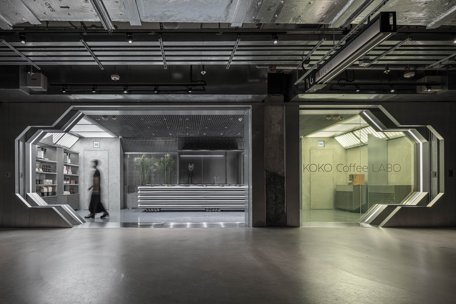 A modern, industrial-style coffee shop with concrete walls, geometric doorways, and a person walking inside. The sign reads “KOKO Coffee LABO.”.
