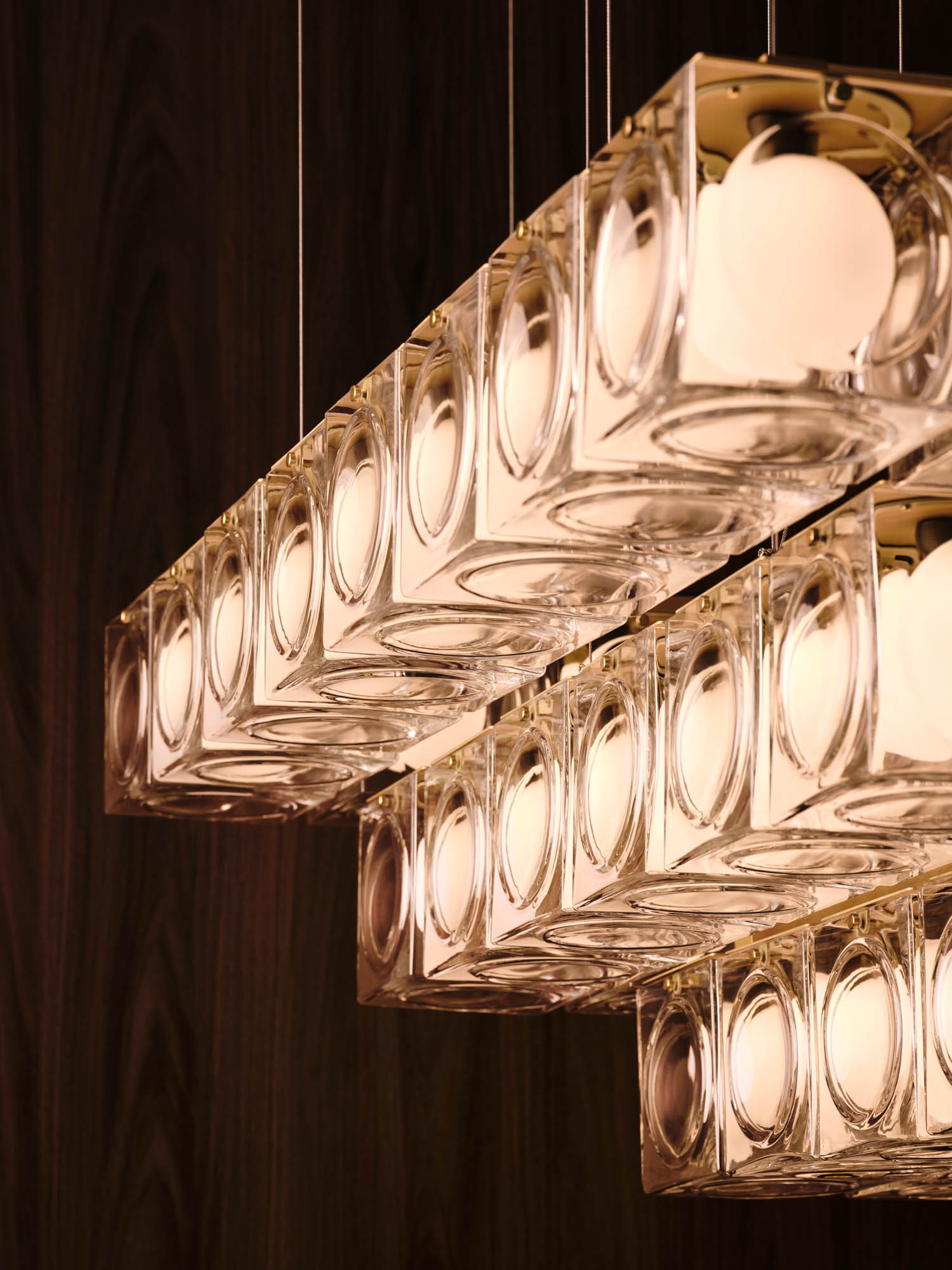 Close-up view of three rectangular, glass pendant light fixtures with round bulbs, suspended in front of a dark wooden wall.