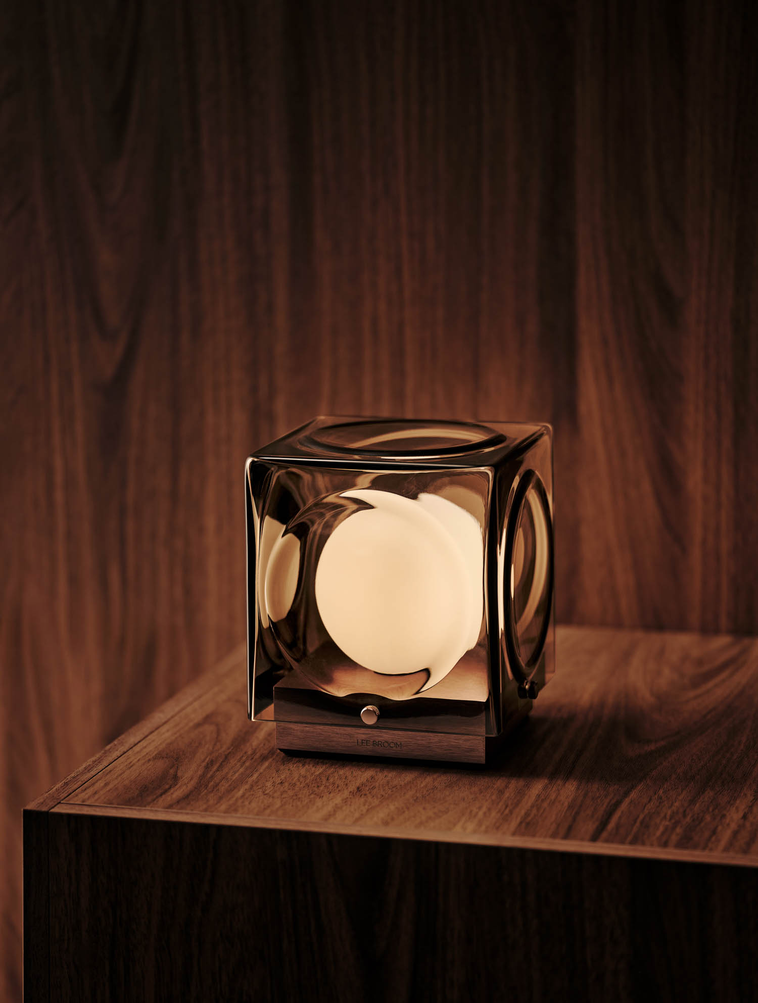 A glass cube lamp with a glowing spherical light inside sits on a wooden surface, against a matching wood background.