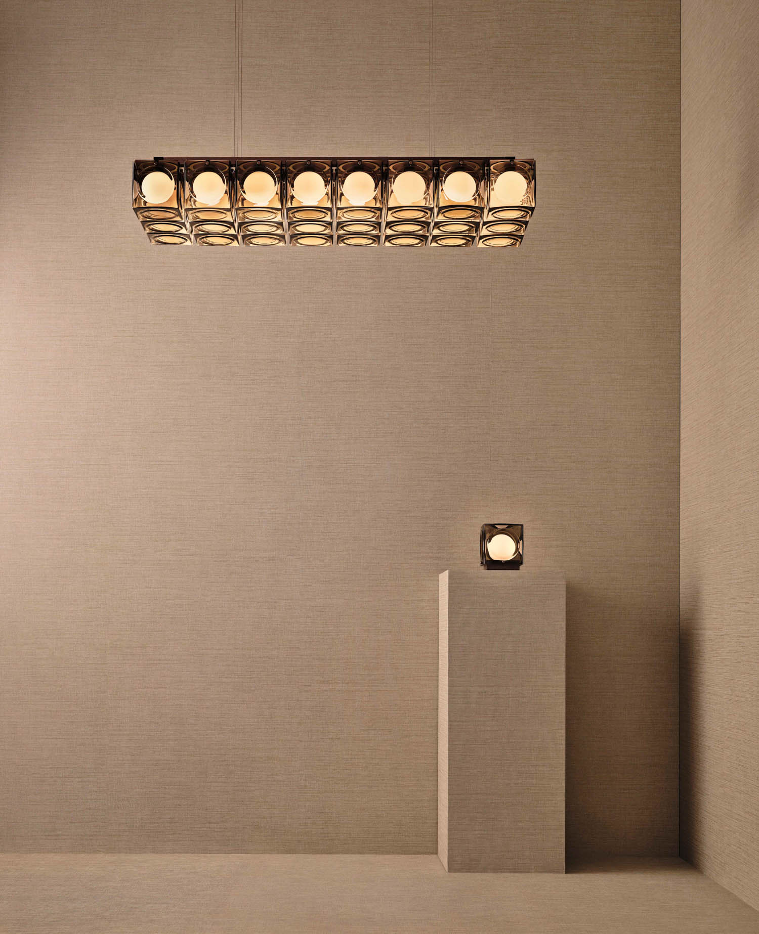 A rectangular pendant light with spherical bulbs hangs above a small sculpture on a pedestal, both in a minimalist beige room.