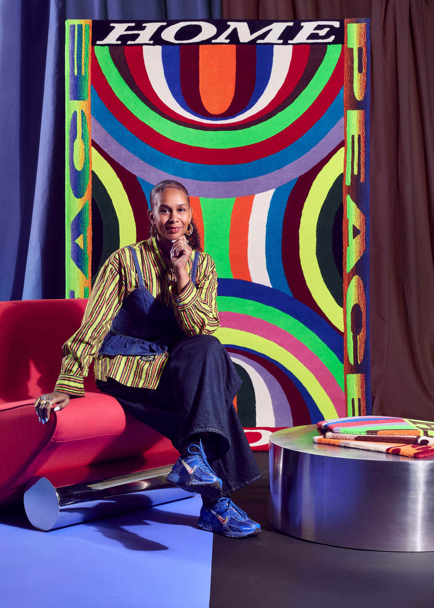 A person sits on a red couch next to a round table, with a colorful backdrop displaying the words “HOME” and “PEACE” in large letters.