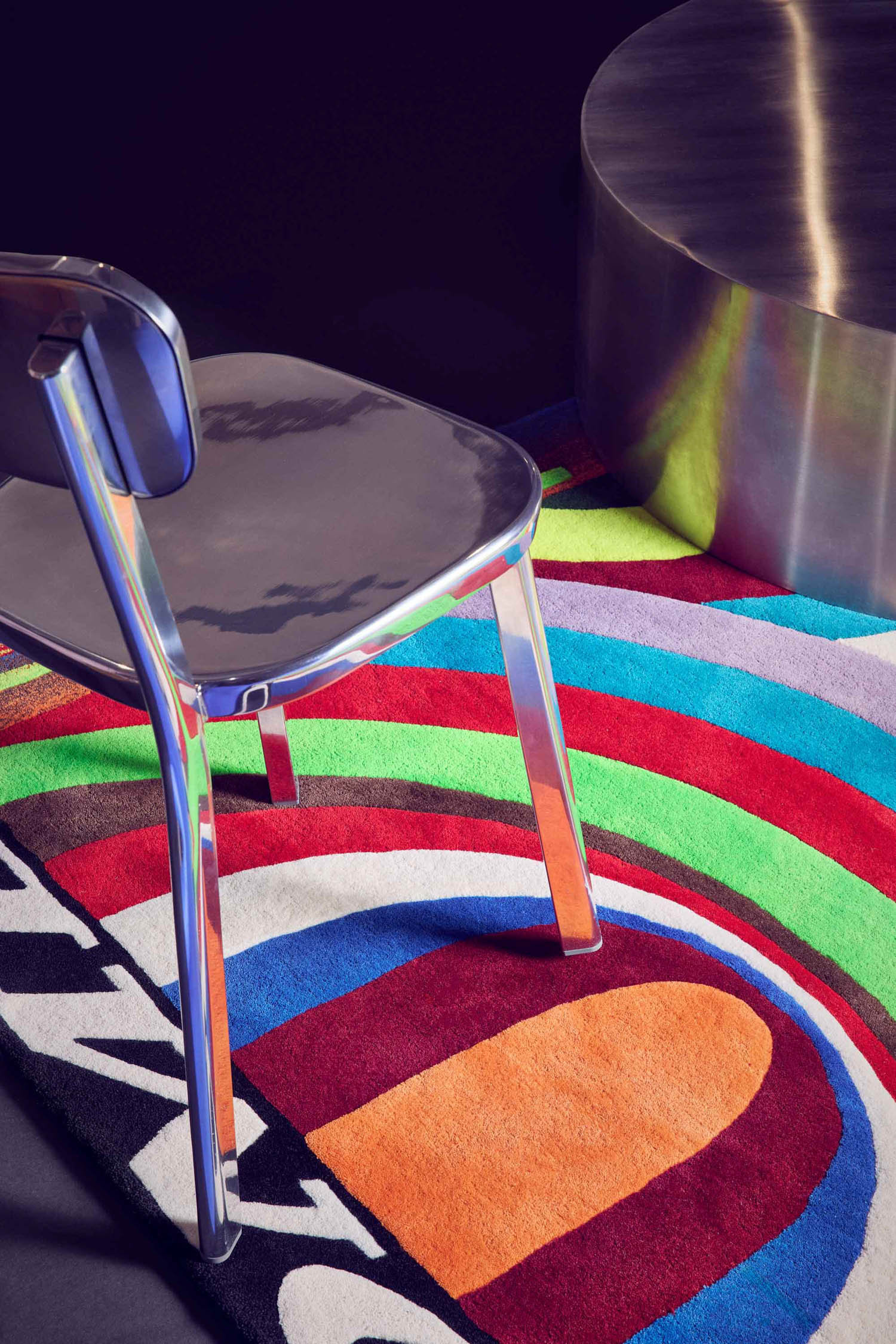 A metallic chair and round table sit on a colorful, abstract-patterned rug with curved shapes and bright shades of red, blue, green, orange, and black.