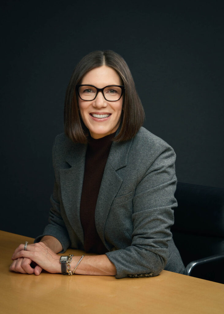A woman with straight brown hair, wearing glasses, a grey blazer, and a dark top, sits at a wooden table and smiles at the camera against a dark background.