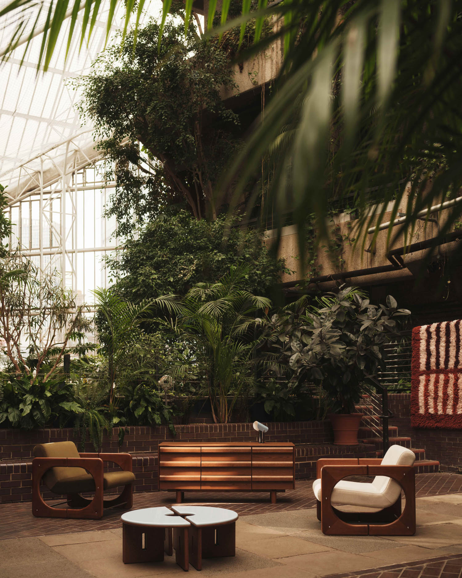 Modern lounge chairs, a wooden sideboard, and round tables are arranged in a lush indoor space with large plants and natural light from a glass ceiling.