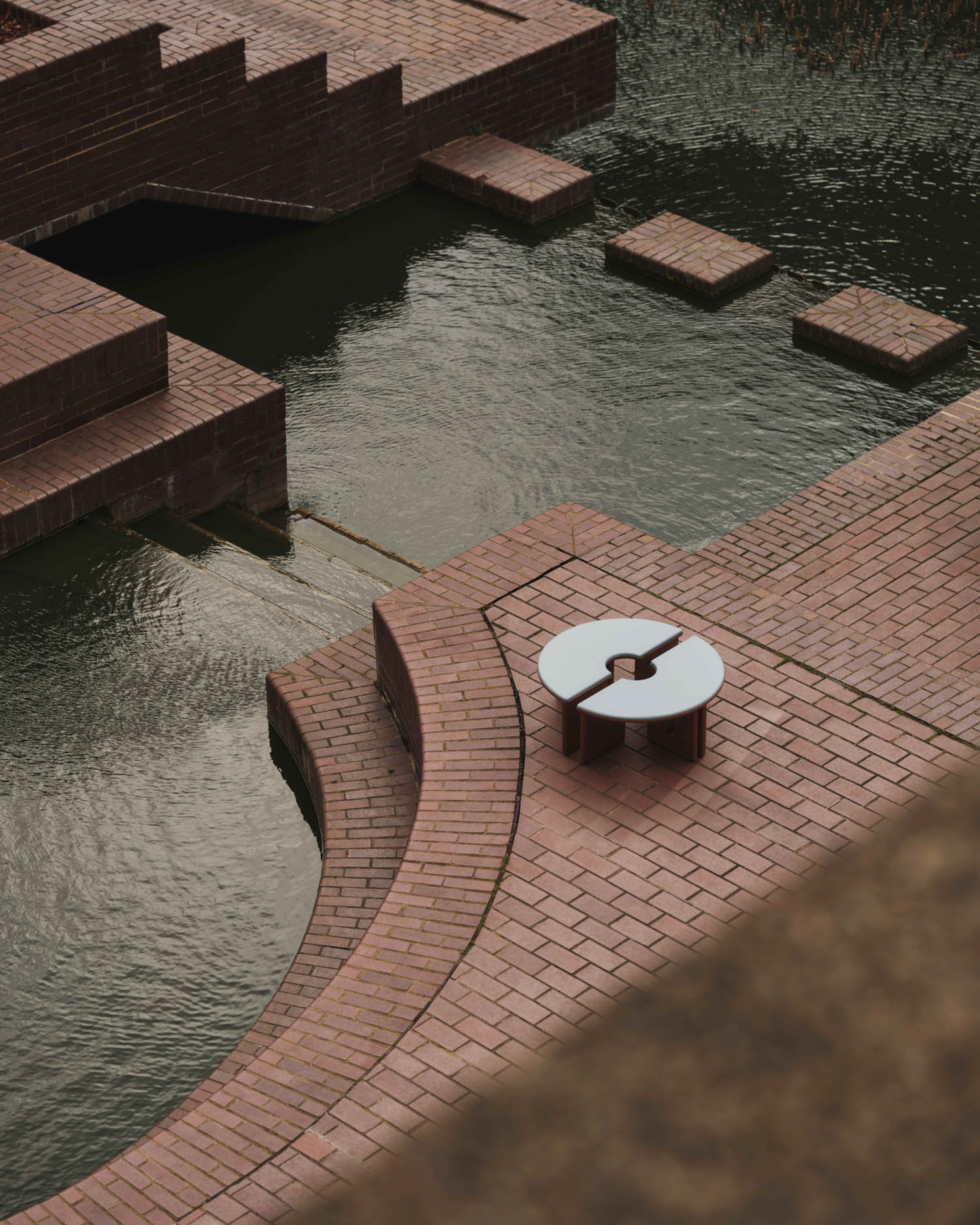 A round white table with a unique cutout design sits on a curved brick patio next to a stepped water feature with brick platforms.