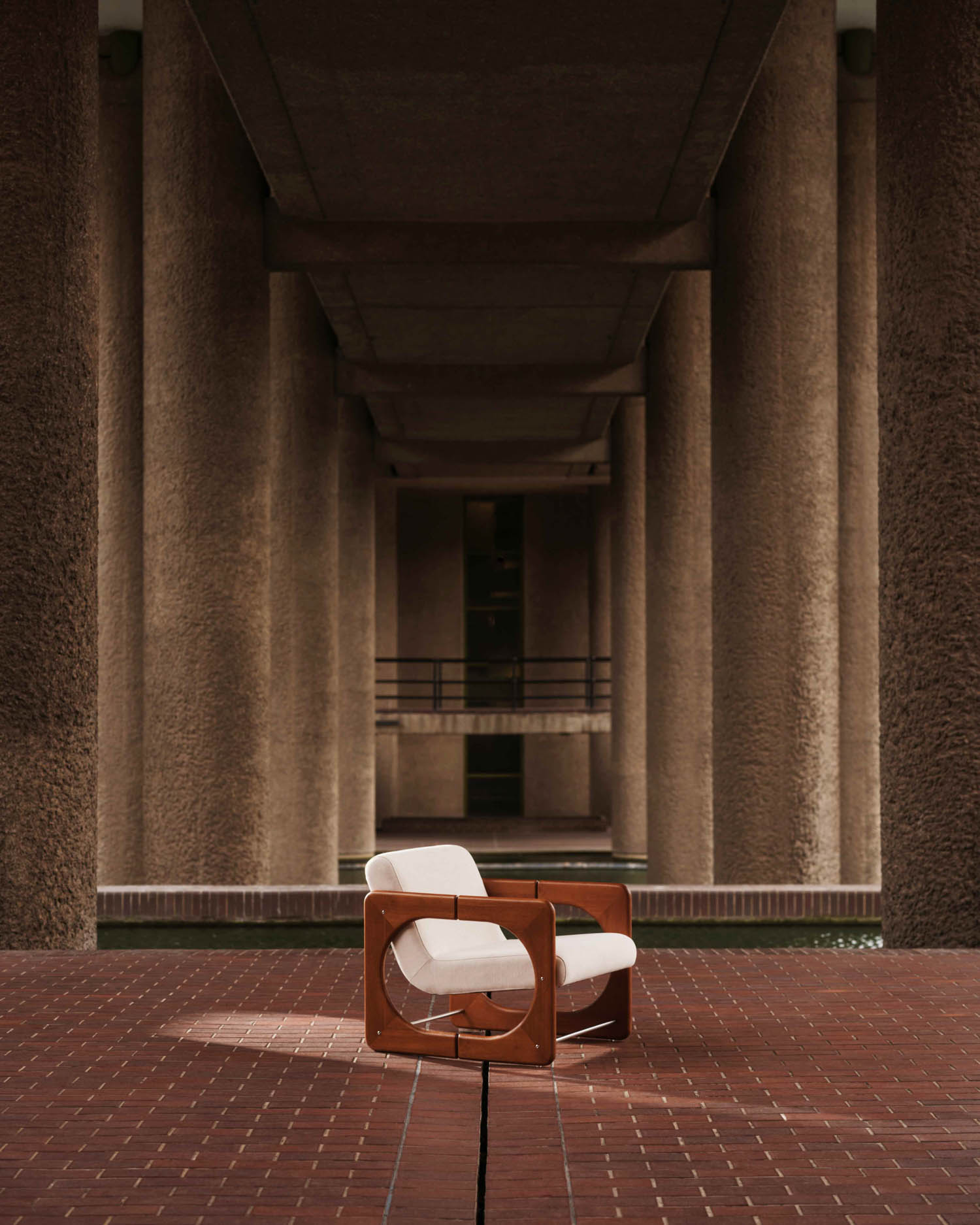A modern wooden armchair with white cushions sits on a red-tiled floor, framed by tall, textured concrete columns in a symmetrical architectural setting.