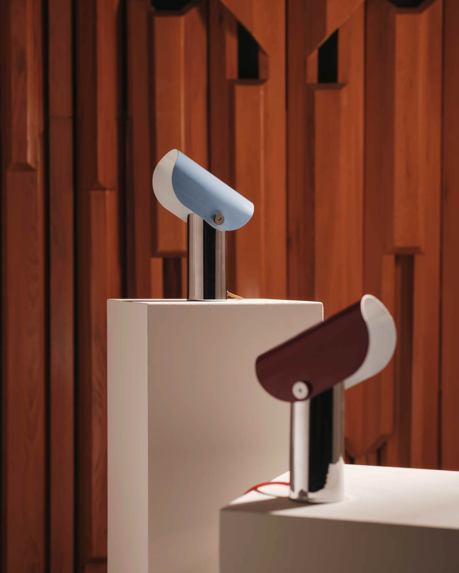 Two modern lamps, one blue and one red, are displayed on white pedestals against a wooden paneled background.