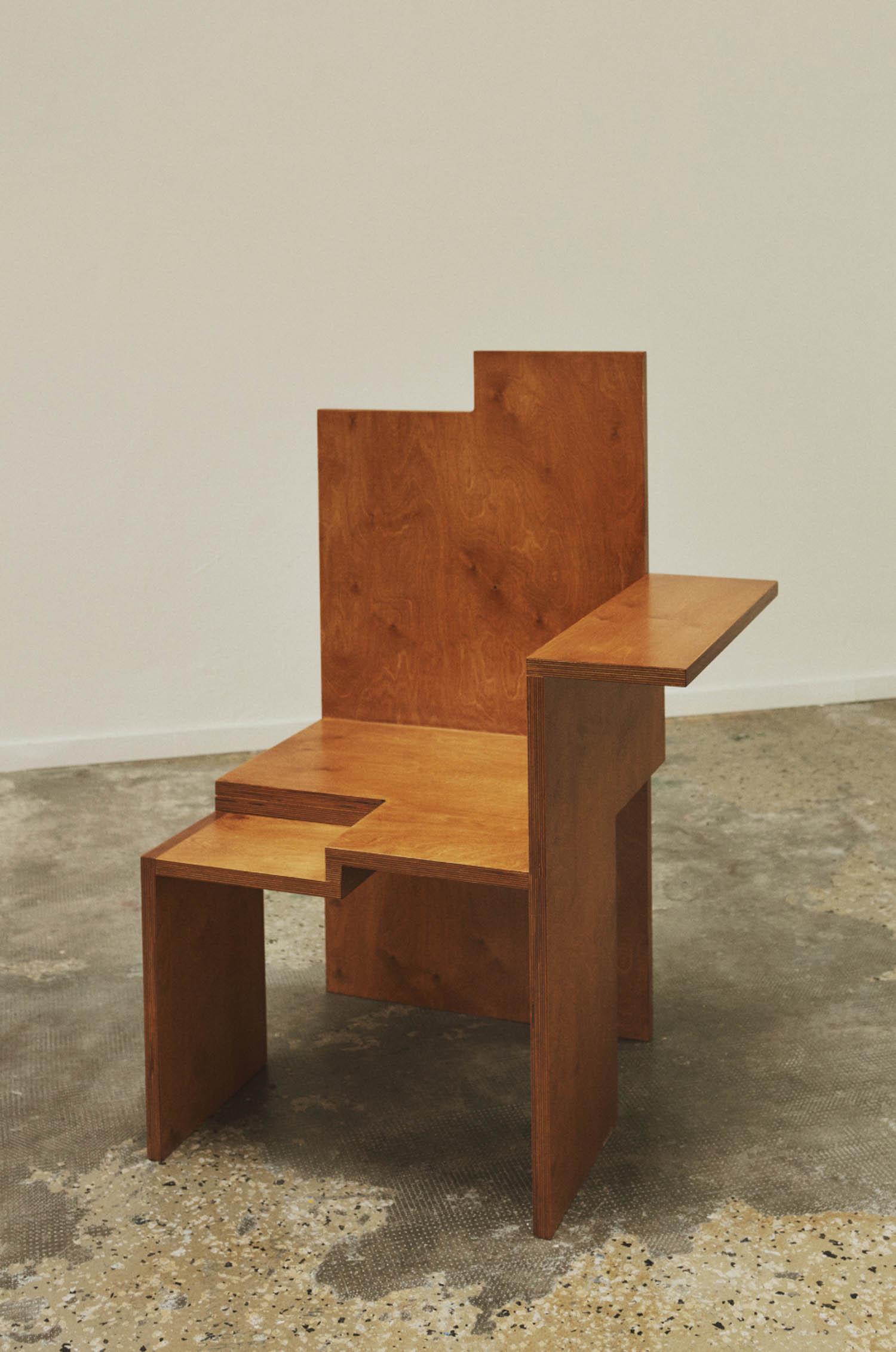 A geometric wooden chair with an angular backrest, integrated side table, and a minimalist design sits on a stained concrete floor against a plain wall.