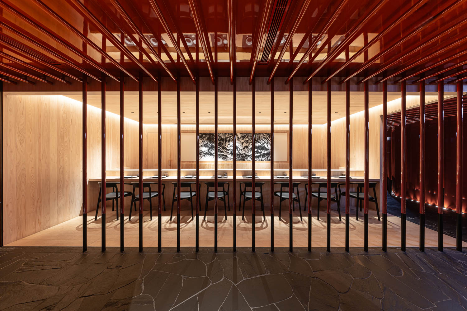 Modern dining room with wooden slats, a long table with chairs, warm lighting, and a black and white wall art centerpiece. Stone flooring is visible in the foreground.