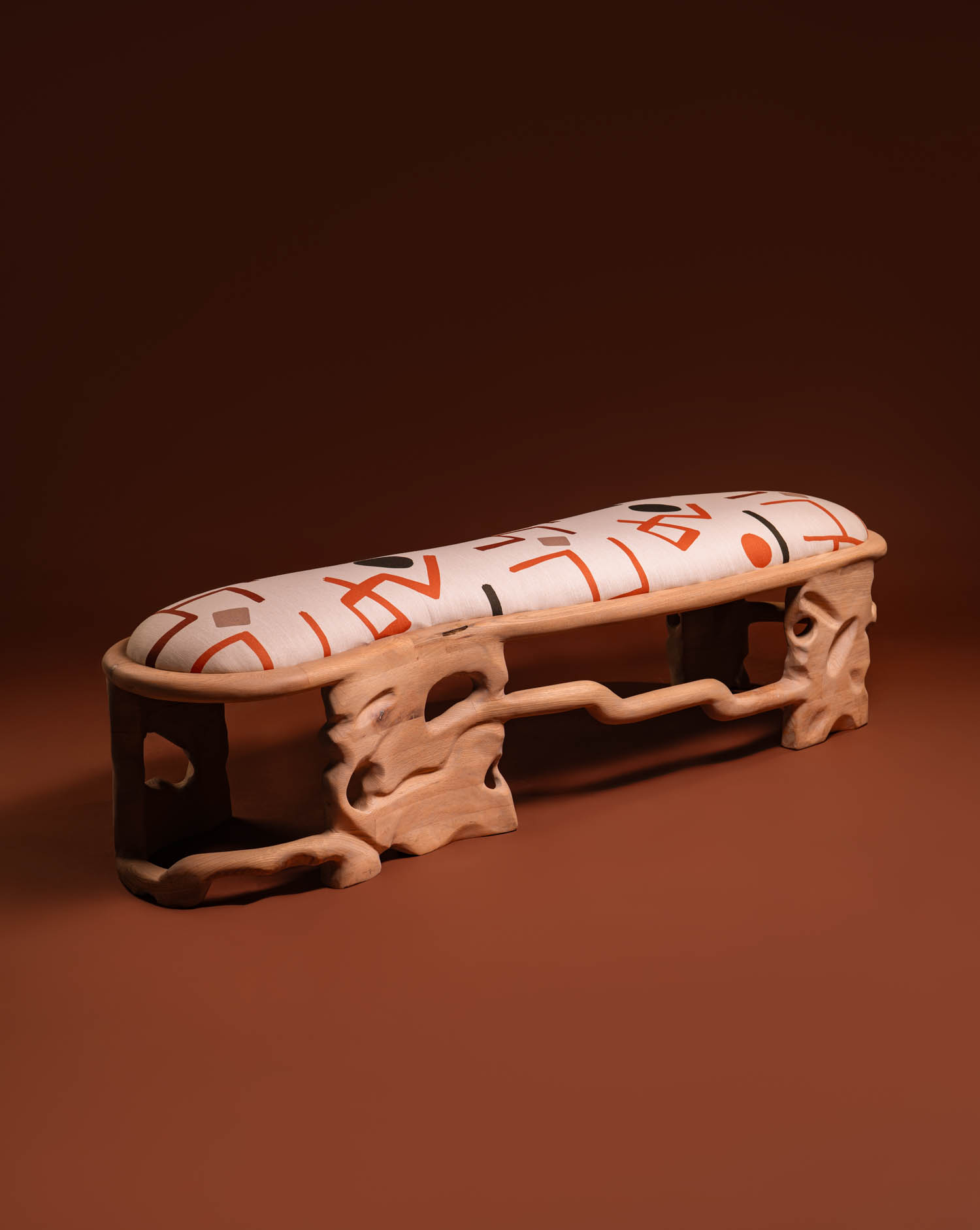 Wooden bench with sculpted, organic legs and a cushioned seat featuring bold, abstract orange and red lettering on a white background, set against a brown backdrop.