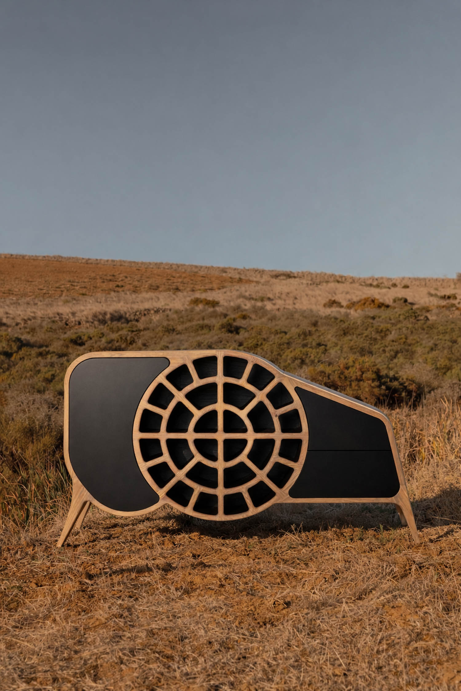 A modern wooden cabinet with circular and rectangular patterns stands on dry grass in a hilly outdoor landscape under a clear sky.