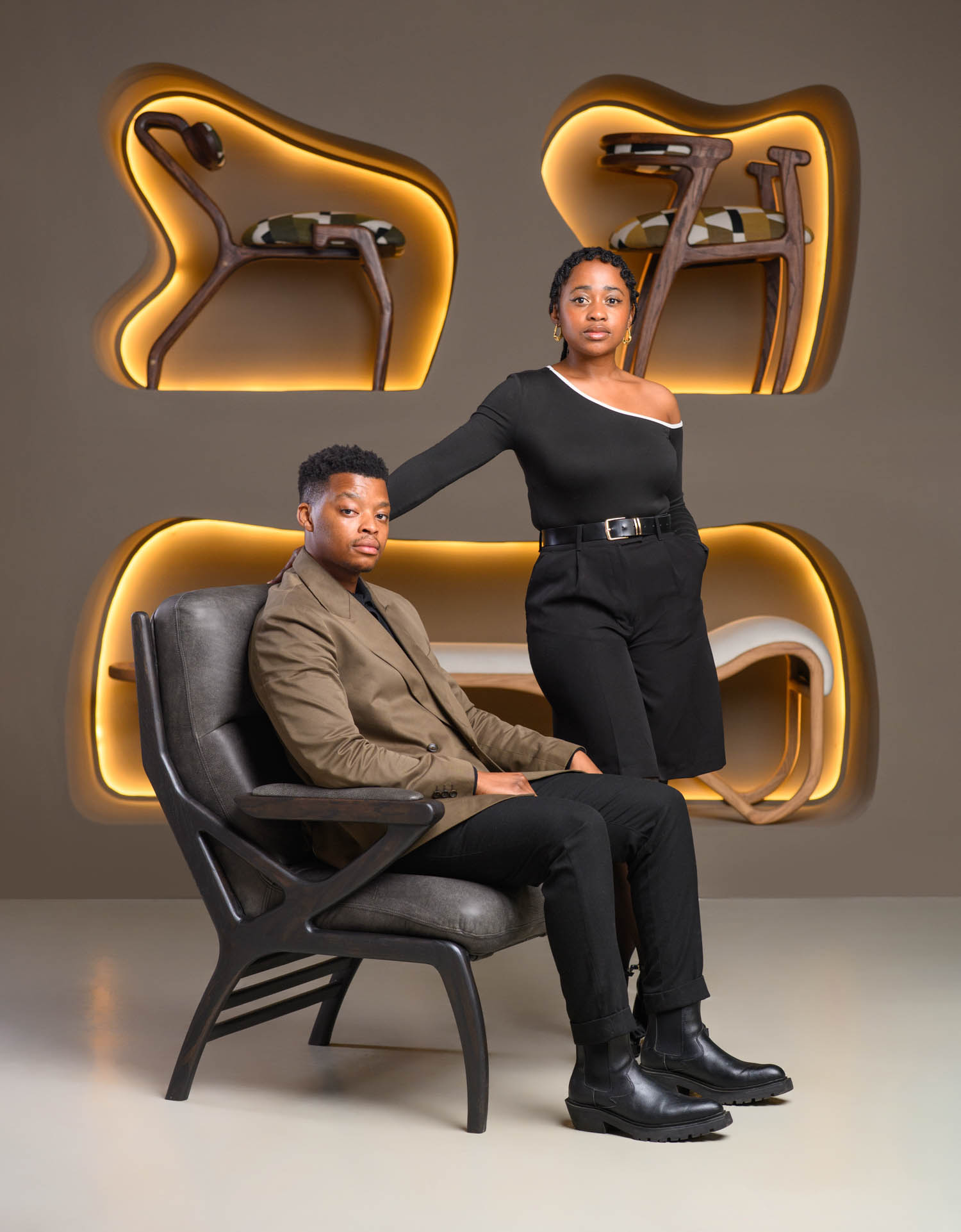 A man sits in a dark chair while a woman stands beside him, both wearing dark clothing, with modern chairs displayed in backlit wall niches behind them.