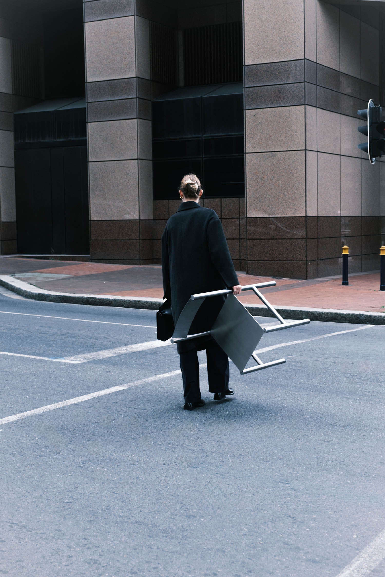 A person in a black coat stands on a city street holding a chair in one hand and a briefcase in the other, with tall office buildings in the background.