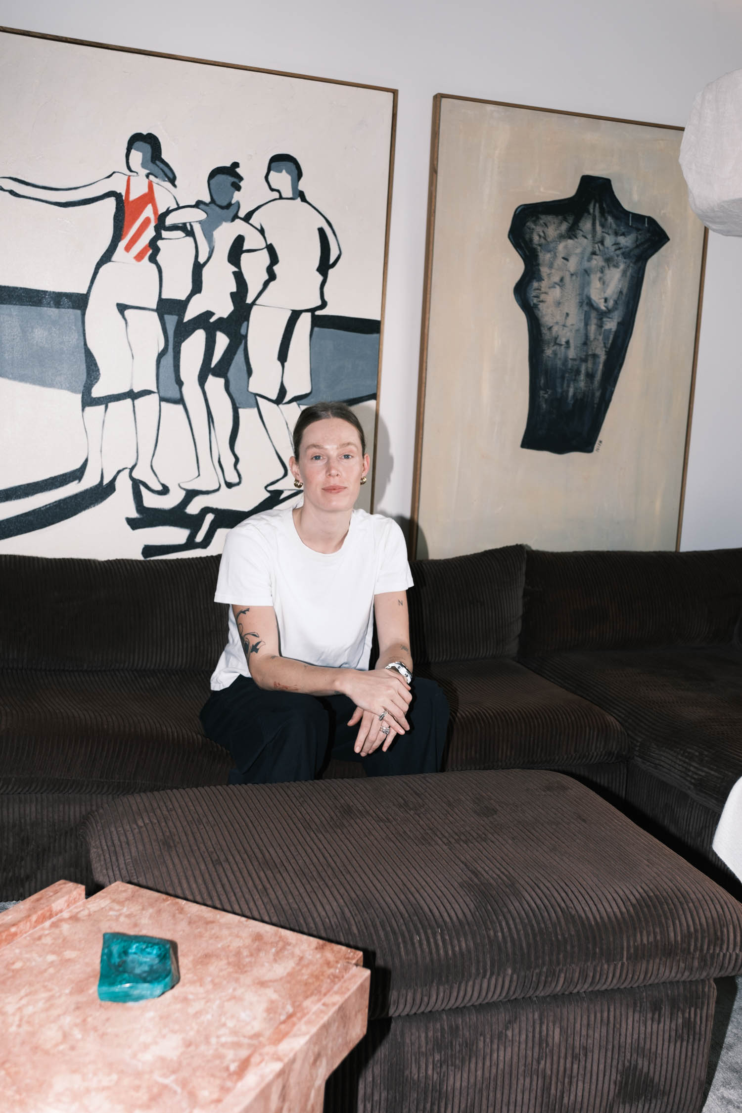 Person in white t-shirt sits on a dark sofa with abstract art on the wall behind them and a pink stone table with a green object in the foreground.
