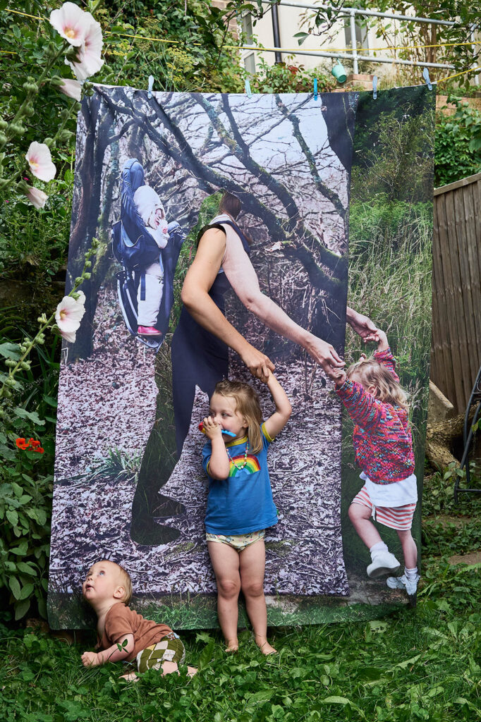 Three children pose in a garden with a large outdoor photo backdrop. The backdrop shows an adult walking with two children, and one child in front aligns with part of the image.