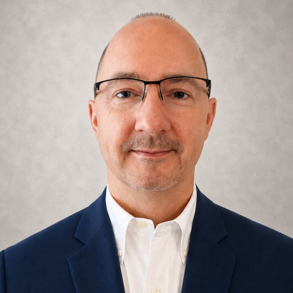 Bald man with glasses wearing a navy suit and white shirt, facing forward against a light gray background.