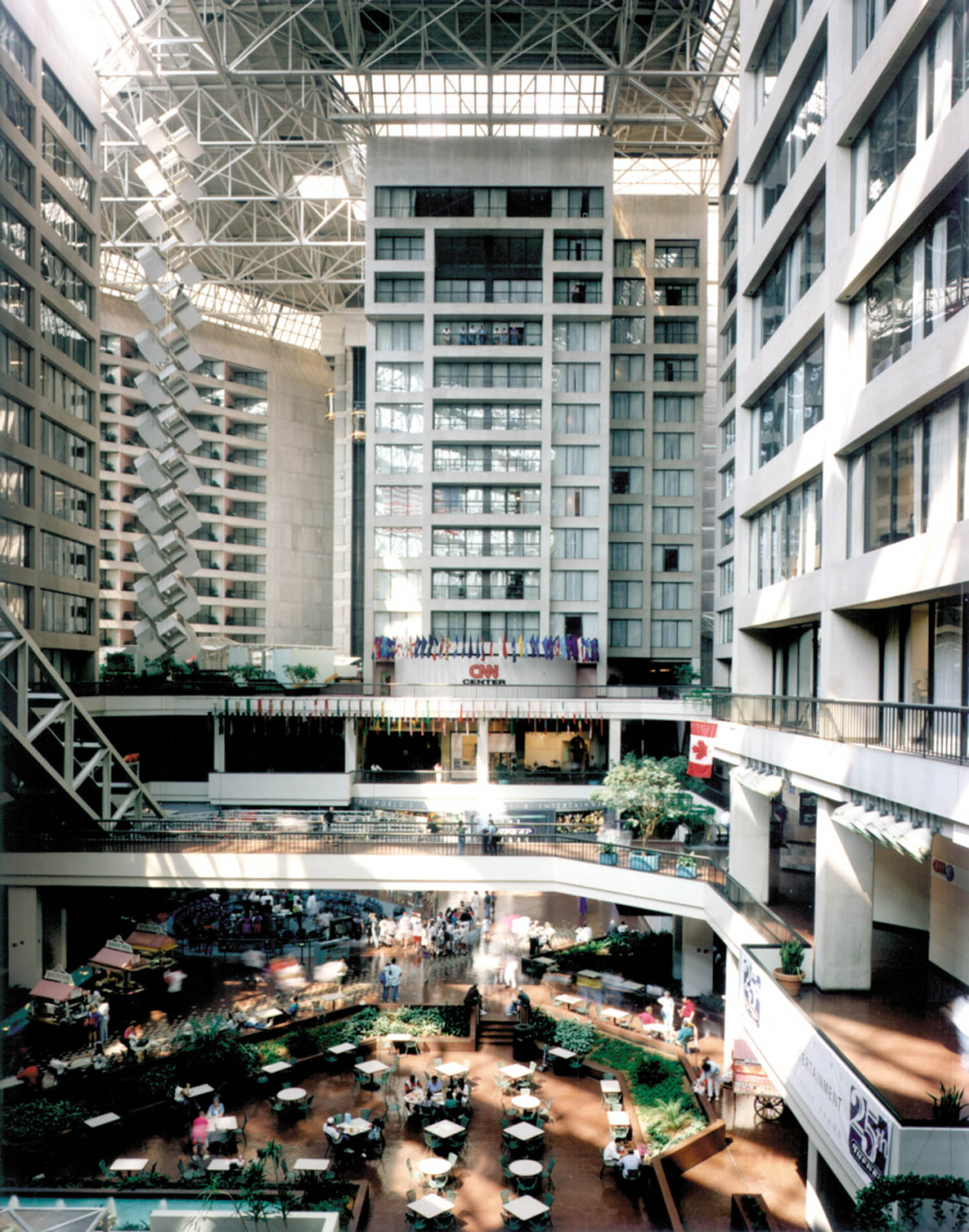 Indoor atrium with tall glass ceiling, multi-story balconies, flags displayed, and people gathered around tables and a garden area on the ground floor.