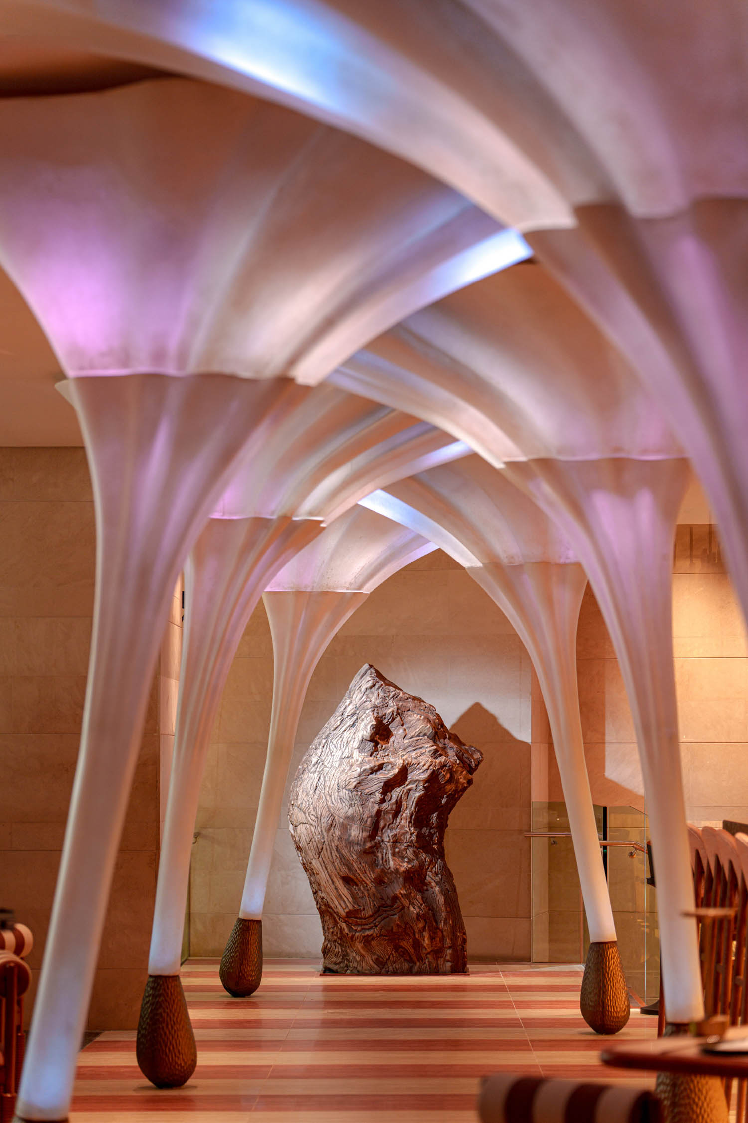 A sculpted wooden structure stands at the end of a hallway lined with white, fluted columns and vaulted ceilings illuminated by soft purple lighting.