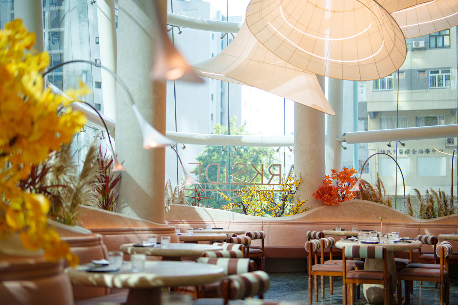 Modern restaurant interior with round tables, cushioned chairs, large windows, yellow and orange floral decor, and oversized white hanging light fixtures.
