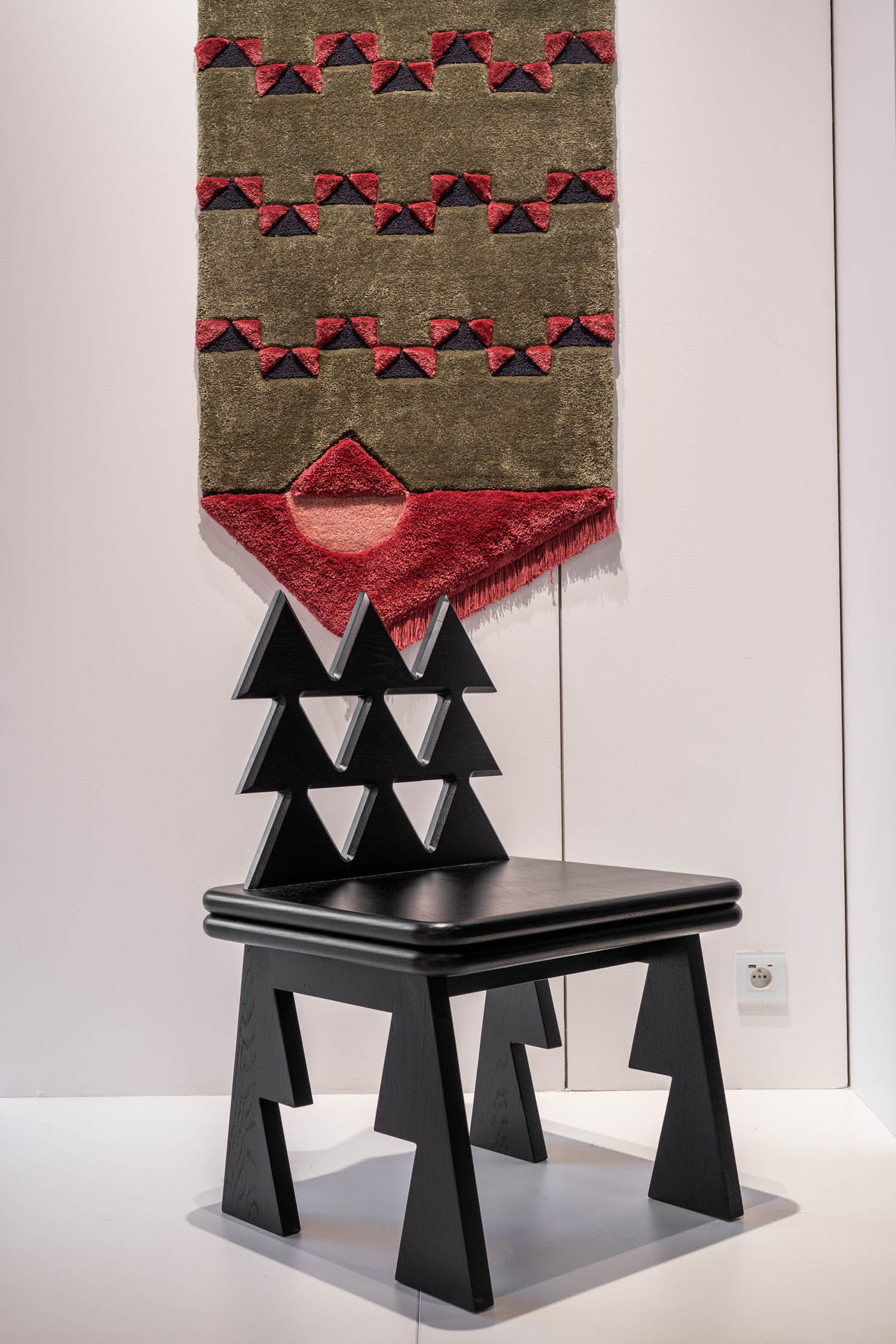 A geometric black wooden chair with zigzag cutouts sits in front of a textured, patterned wall hanging with red and olive green shapes.