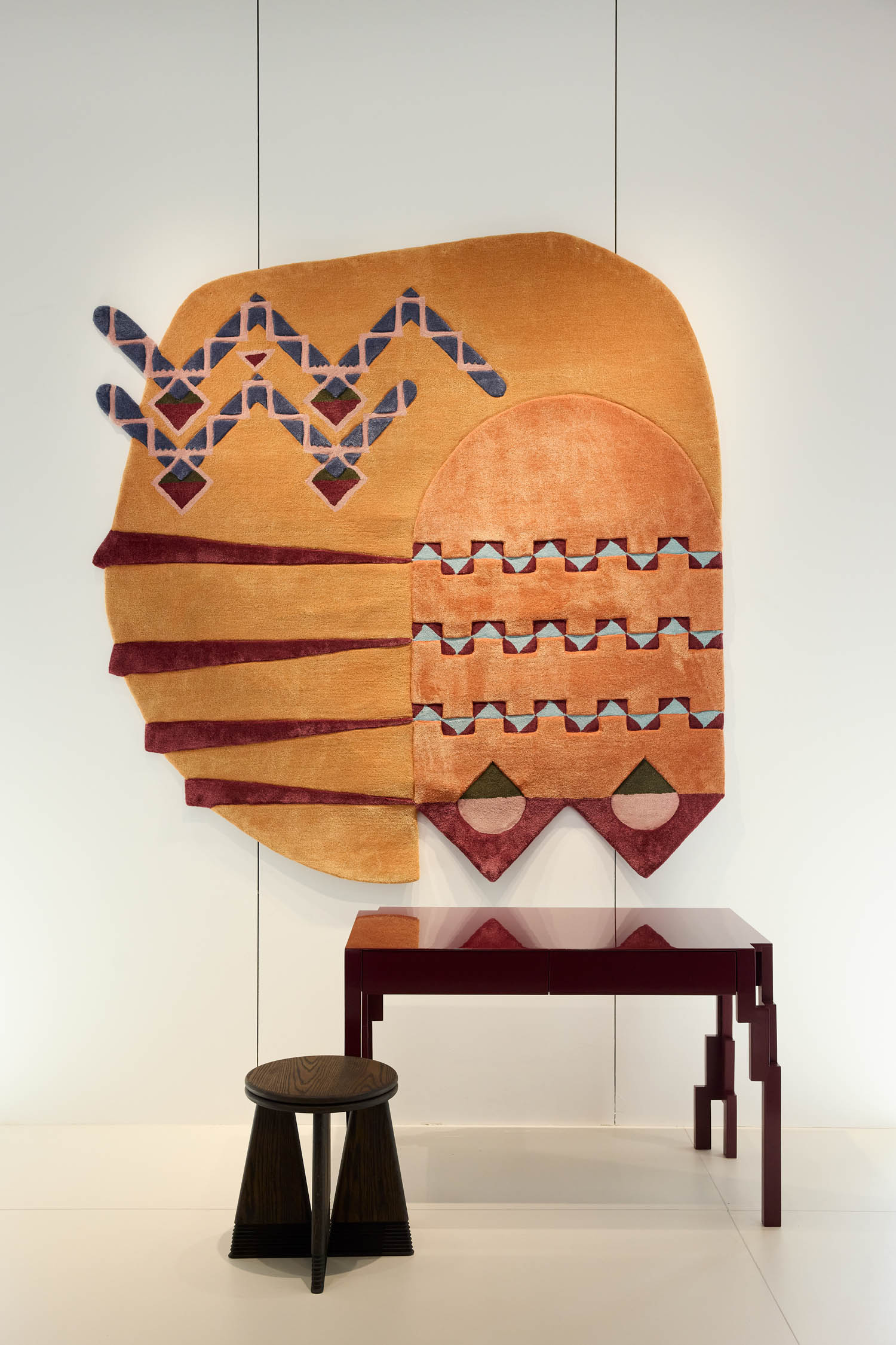 Abstract wall textile with geometric shapes in gold, red, and blue above a red angular desk and a small black stool in a minimalist white space.