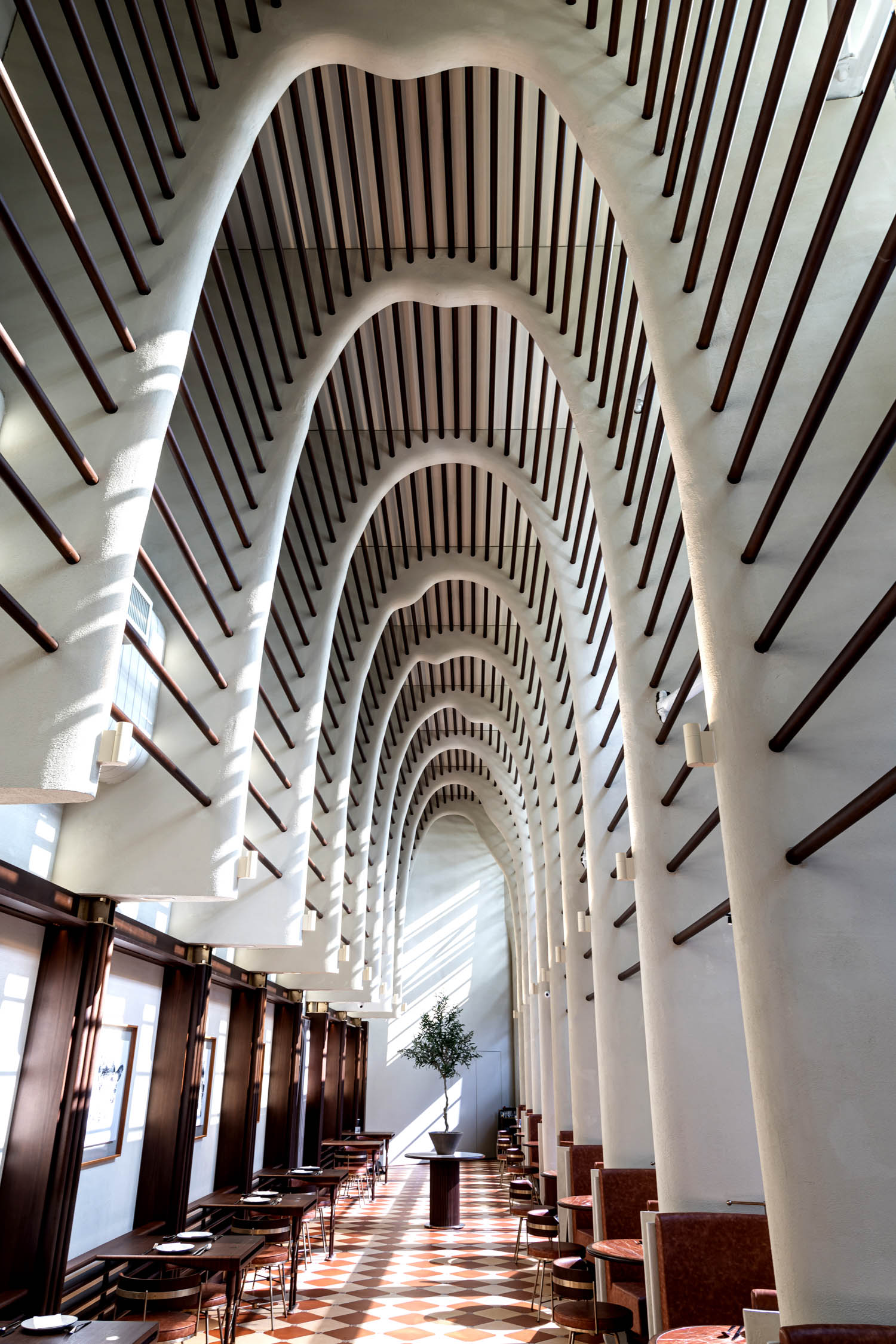 A tall, arched hallway with repeating white columns and wooden beams, lined with tables and chairs along one side, and sunlight streaming in through high windows.