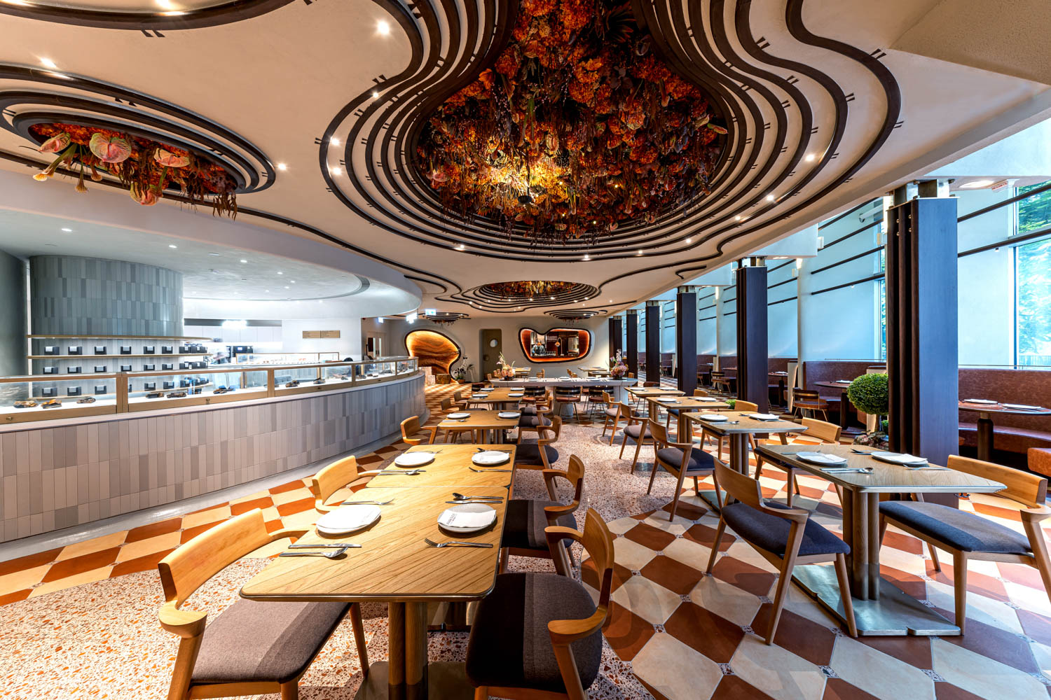 Modern restaurant interior with wooden tables, cushioned chairs, large windows, patterned floor, and an ornate ceiling featuring colorful decor. The space is well-lit and neatly arranged.
