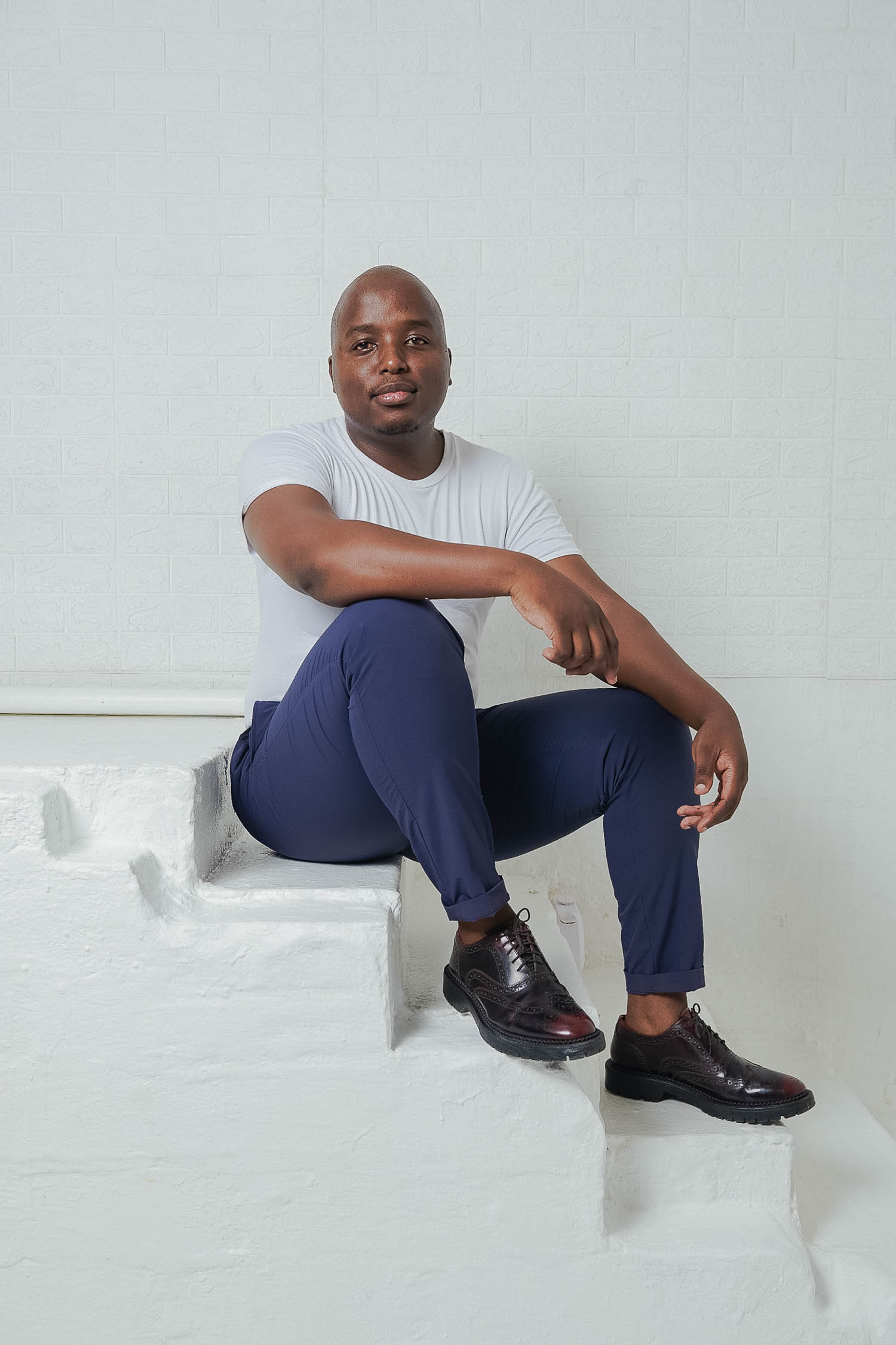 A man wearing a white t-shirt, blue pants, and black shoes sits on white steps against a white brick wall, looking at the camera.