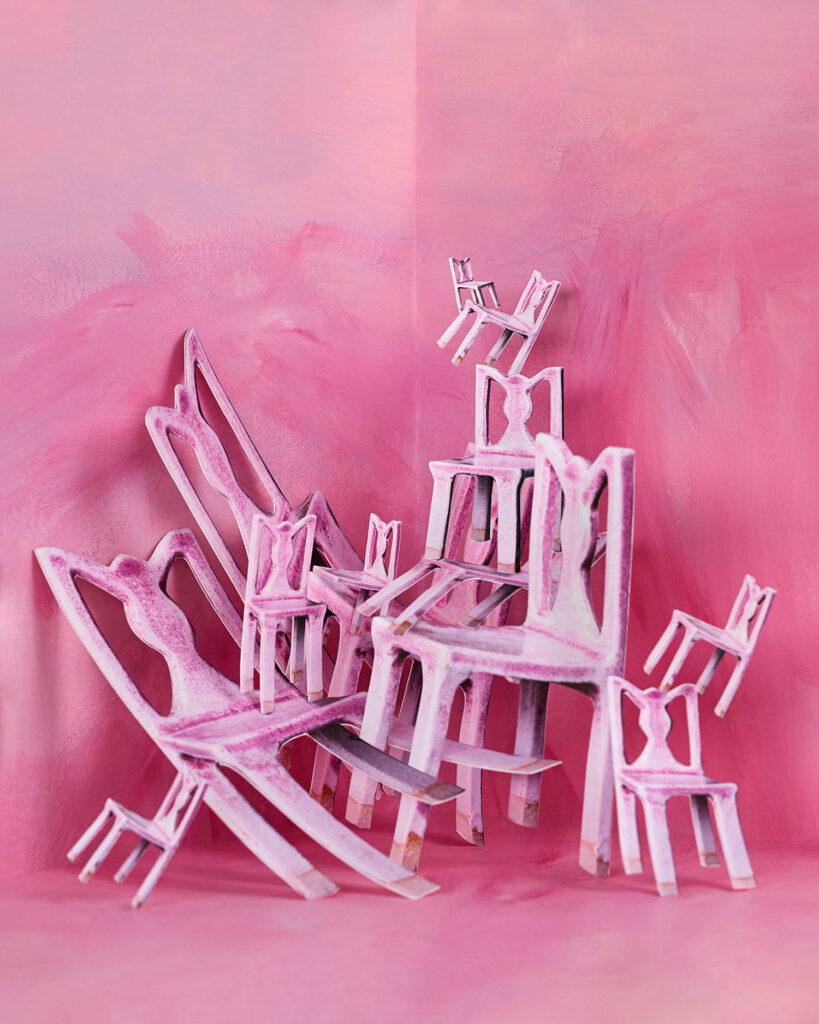 Several pink, paper cutout chairs of varying sizes are arranged in a scattered, chaotic manner against a pink painted background.