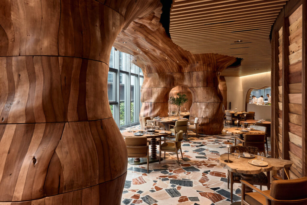 Restaurant interior with sculpted wooden columns, large windows, modern furniture, and a patterned tile floor.