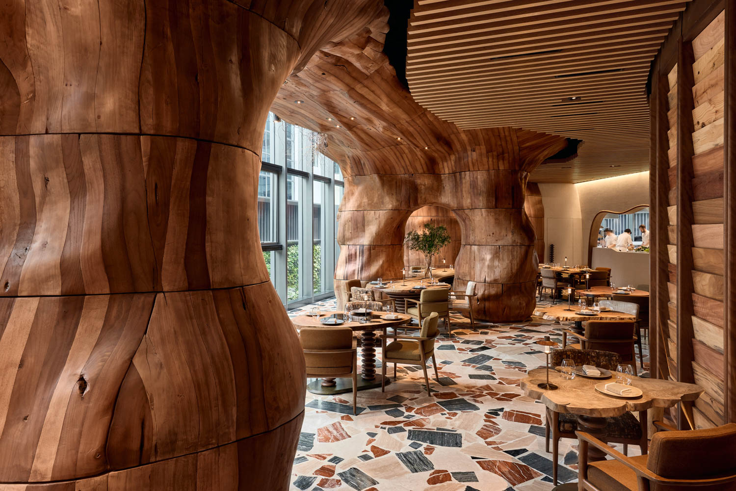 Restaurant interior with sculpted wooden columns, large windows, modern furniture, and a patterned tile floor.