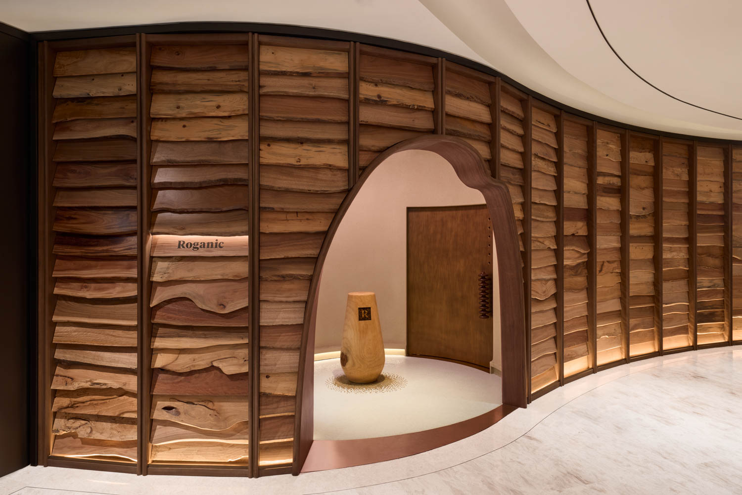 Curved wooden wall with a wavy doorway entrance labeled “Roganic”; smooth wood textures and minimalist decor inside.