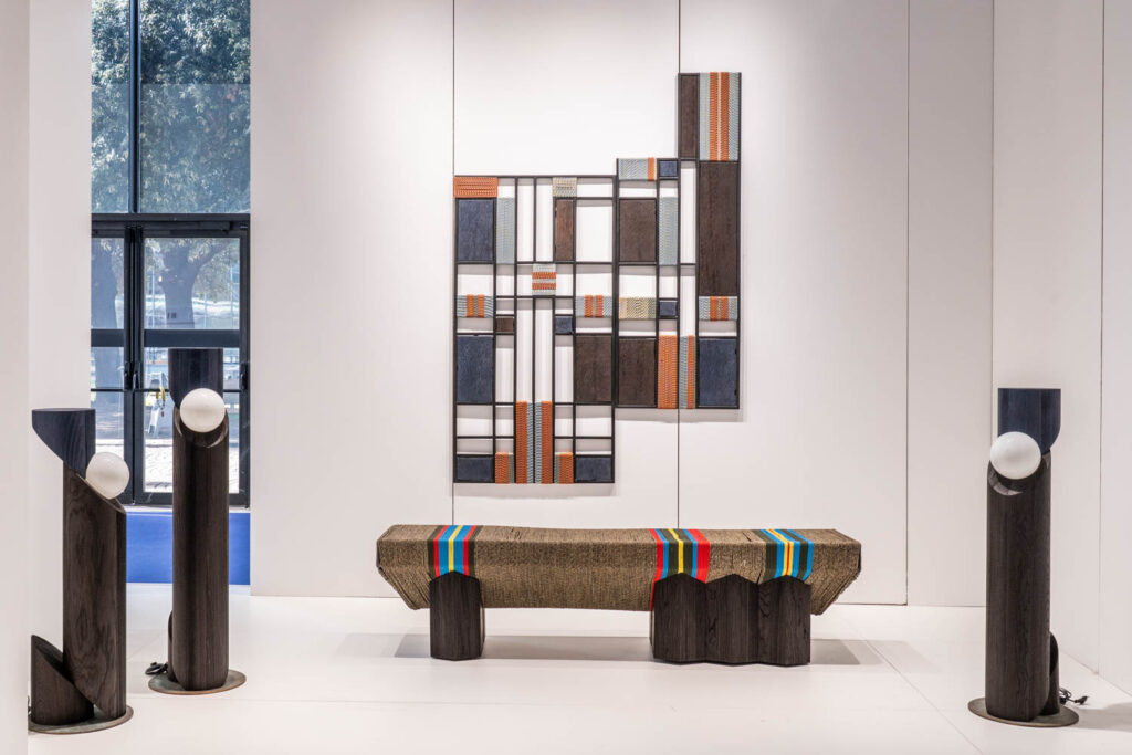 A modern art installation featuring a geometric wall piece, a textured bench with colorful stripes, and four dark pillars topped with round white lights.
