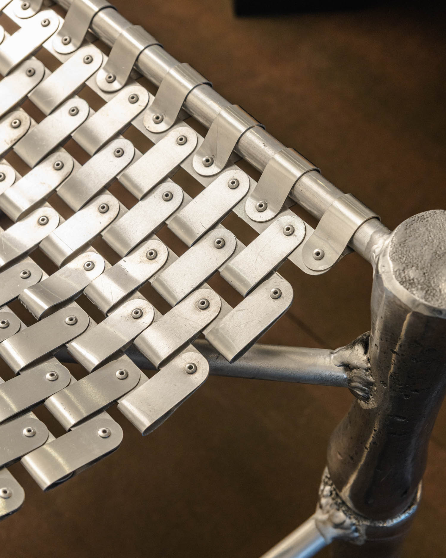 Close-up of a metal chair showing interwoven aluminum strips fastened with rivets, creating a grid pattern on the seat and backrest.