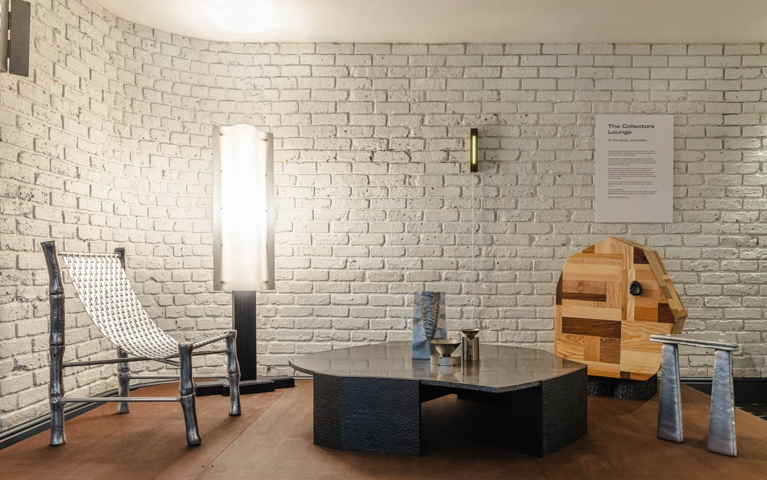 Modern lounge area with a white brick wall, unique metal and wood furniture, woven chair, geometric table, and wall-mounted light fixtures. A sign is displayed on the wall.