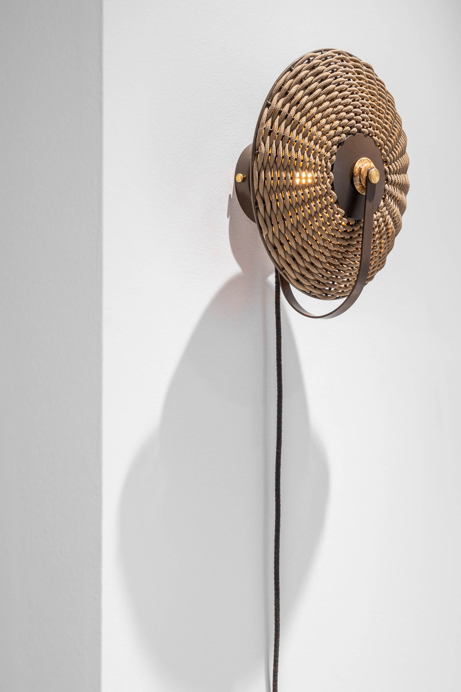 Round wicker wall sconce with a black cord, mounted on a white wall, casting a distinct shadow to the left.