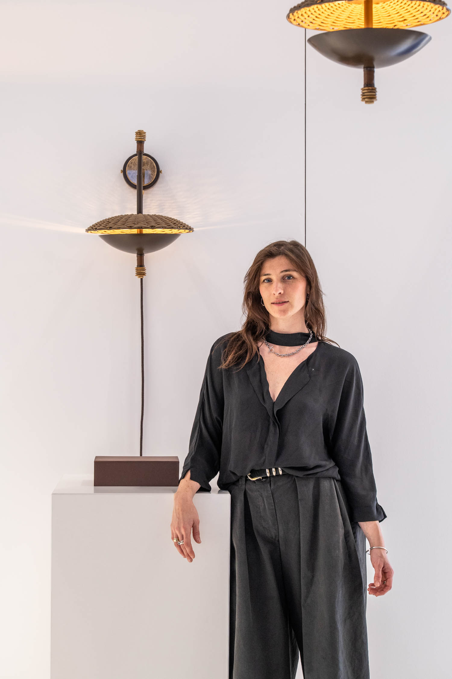 Woman in loose black clothing stands beside a white pedestal with wall and ceiling lights above, against a plain white background.