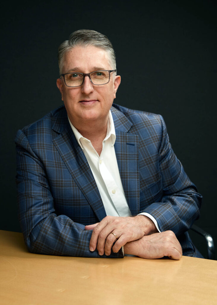 A man with gray hair and glasses wearing a blue plaid blazer and white shirt sits at a wooden table against a dark background.