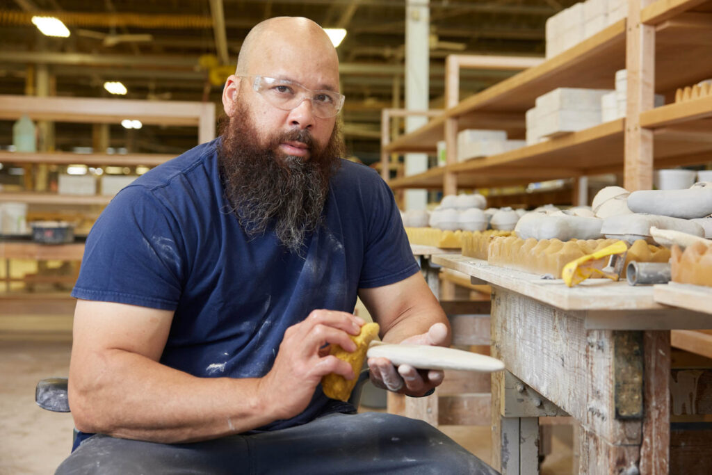 Man with a beard wearing safety glasses sanding a ceramic plate in a pottery workshop, surrounded by shelves with pottery items.