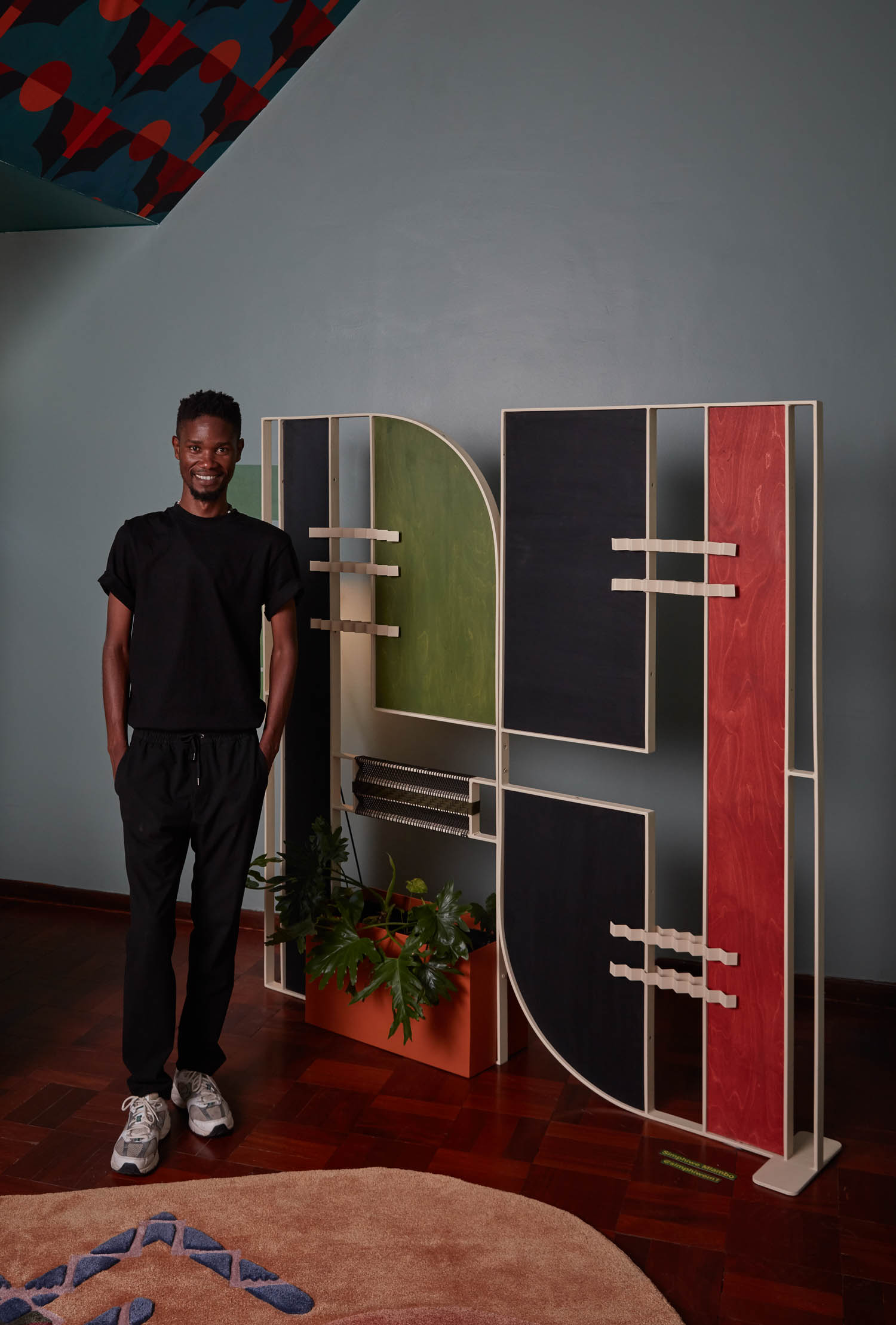 A man in black clothing stands next to a modern, colorful shelving unit with plants on a wooden floor in a room with muted blue walls.