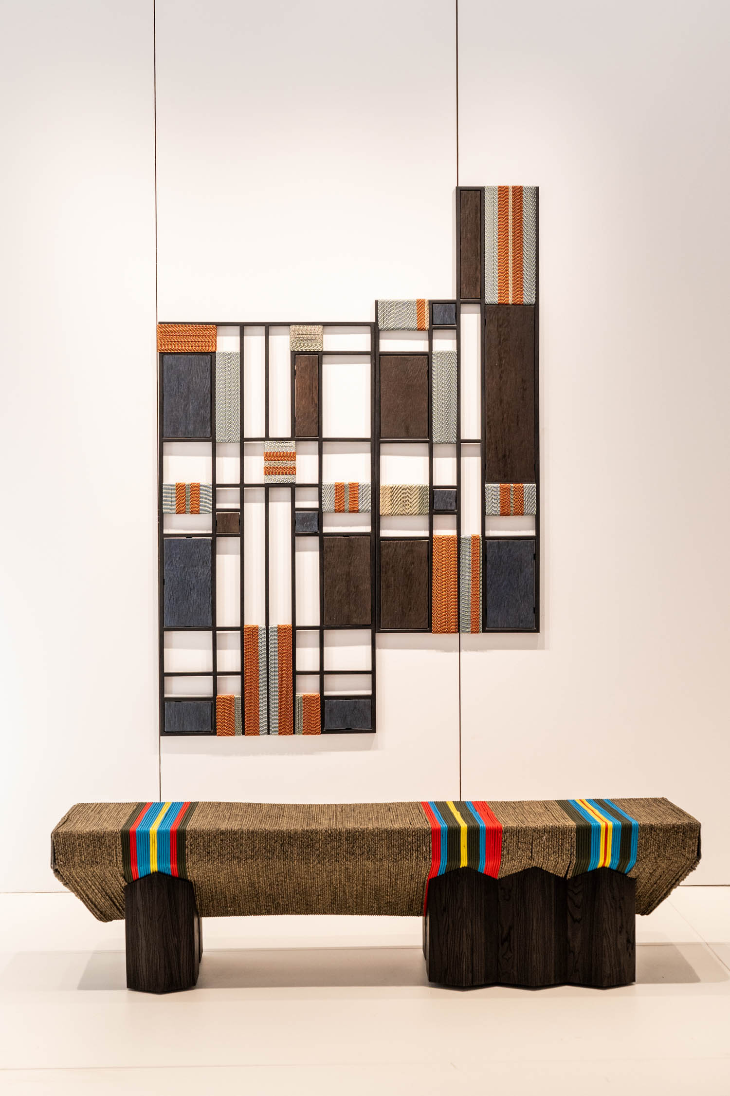 A textured wooden bench with colorful stripes sits in front of a geometric wall art piece made of wood, metal, and patterned panels, displayed against a white background.