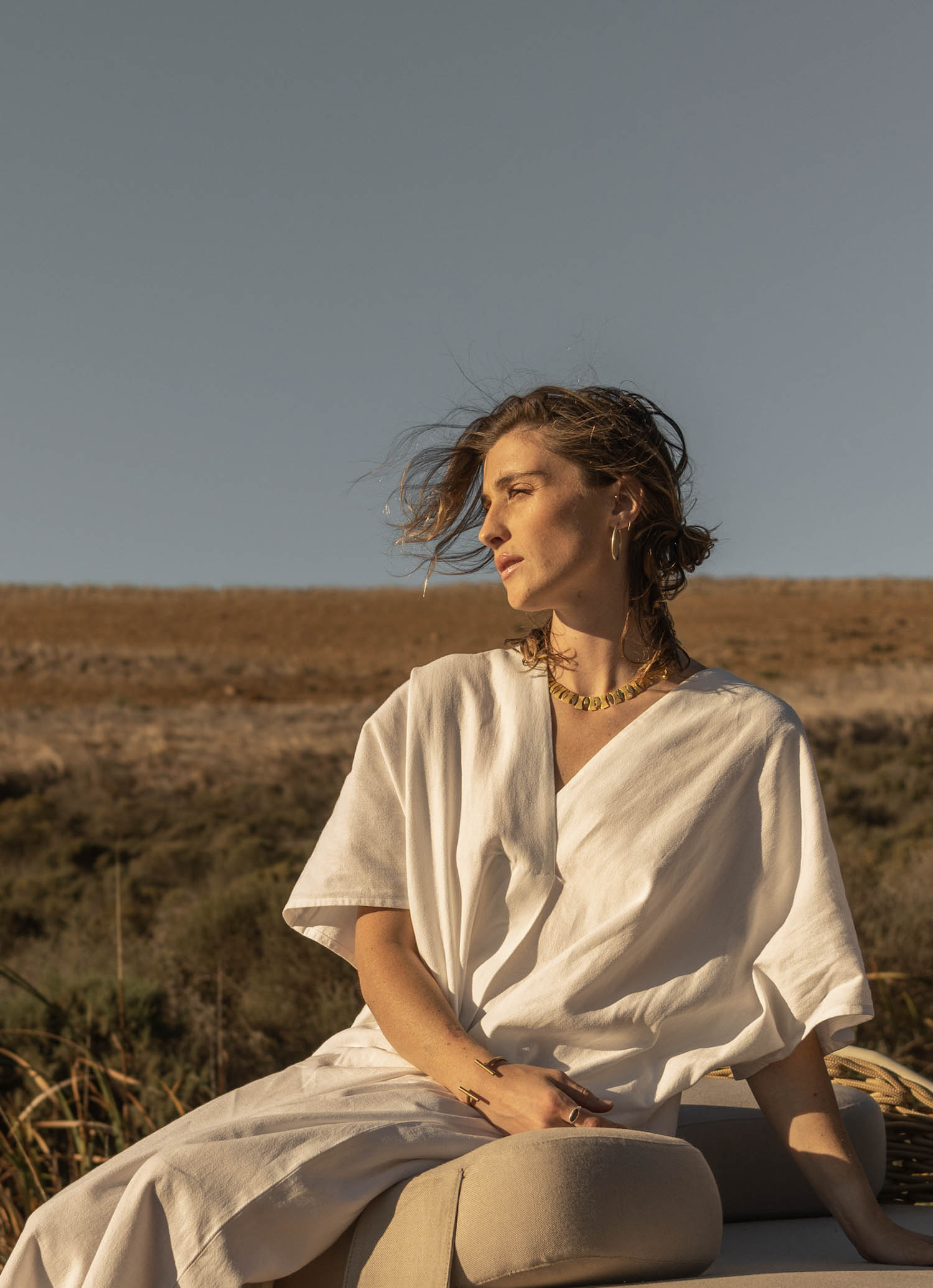 A person in a white robe sits outdoors on a beige cushion, looking to the side, with dry grass and a clear sky in the background.
