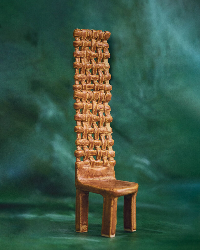 A miniature chair with a tall, woven backrest and four straight legs, displayed against a dark green background.