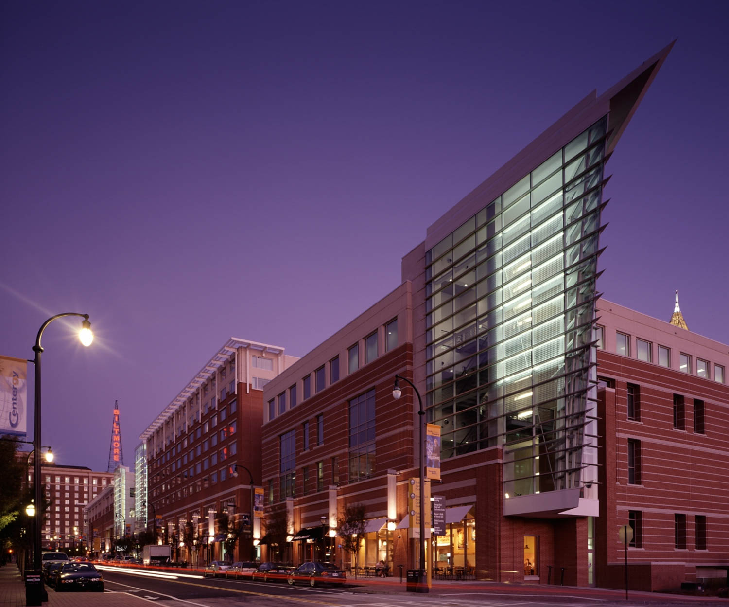 Modern brick building with a sharp angled glass facade, illuminated at dusk, alongside a city street with light poles and cars.