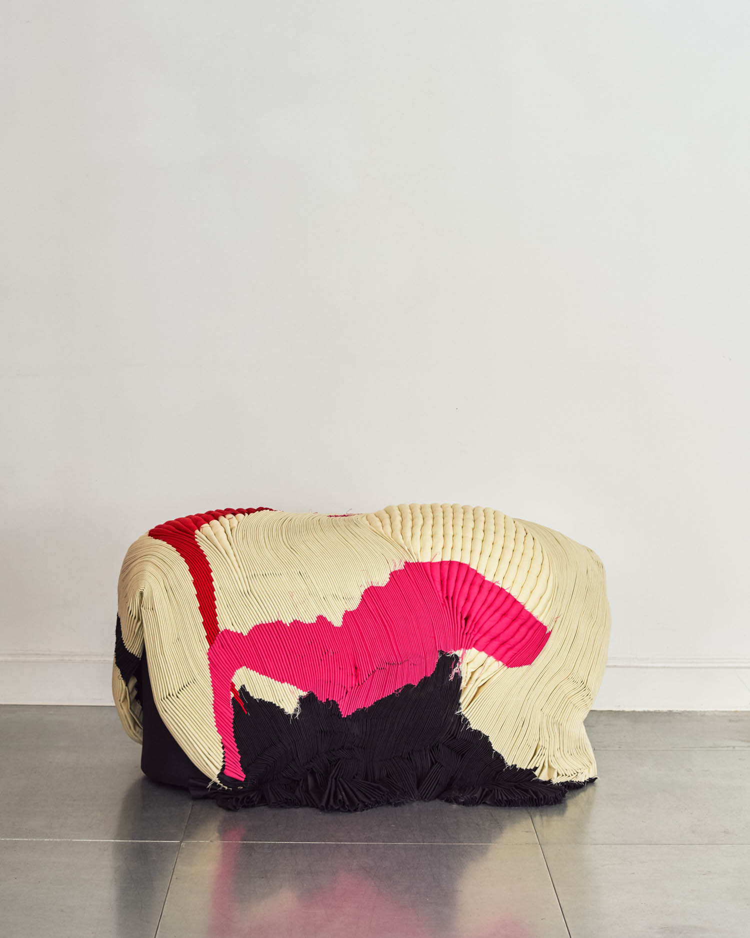 An abstract-shaped object covered with textured fabric in cream, black, and bright pink sits on a gray floor against a plain white wall.